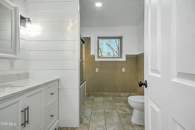 full bath with toilet, vanity, wainscoting, enclosed tub / shower combo, and tile walls