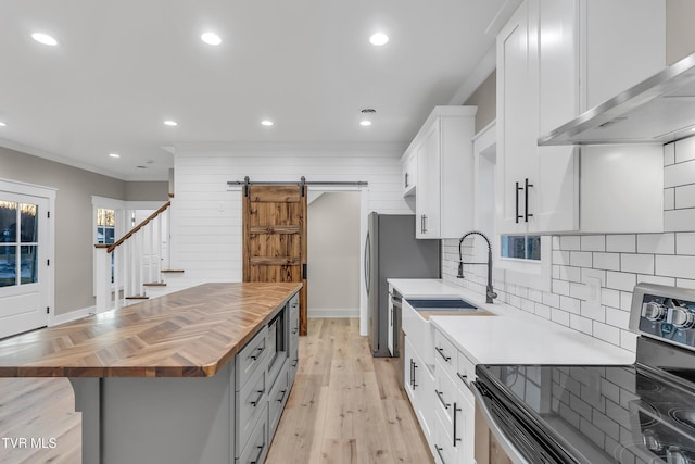 kitchen with wall chimney exhaust hood, a barn door, wooden counters, white cabinets, and appliances with stainless steel finishes