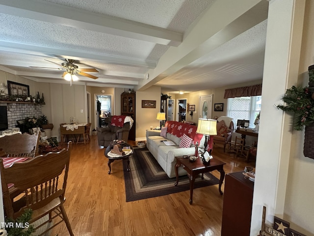 living room with a brick fireplace, ceiling fan, light wood-type flooring, a textured ceiling, and beamed ceiling