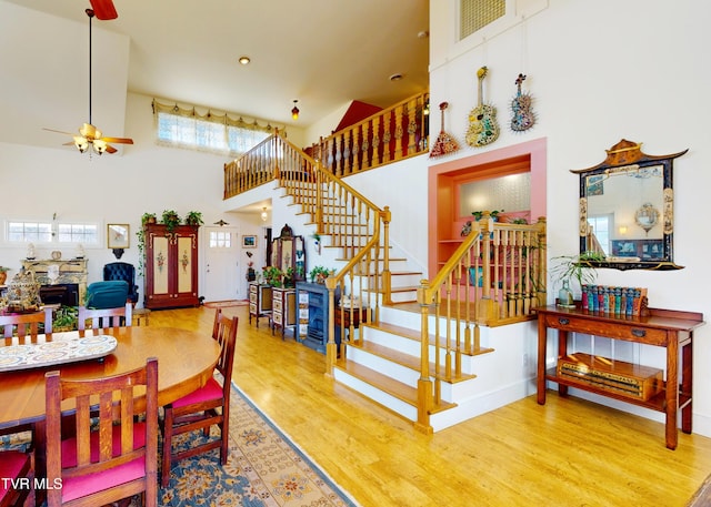 dining space with stairway, a towering ceiling, a ceiling fan, a stone fireplace, and wood finished floors