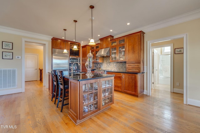 kitchen featuring sink, wall chimney exhaust hood, backsplash, built in appliances, and an island with sink