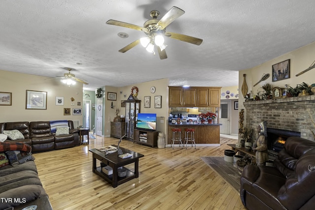 living room with a brick fireplace, ceiling fan, a textured ceiling, and light wood-type flooring