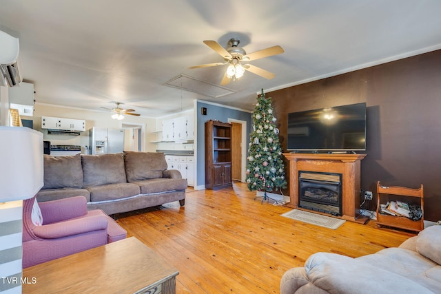 living room with attic access, a ceiling fan, a glass covered fireplace, a wall unit AC, and light wood-type flooring