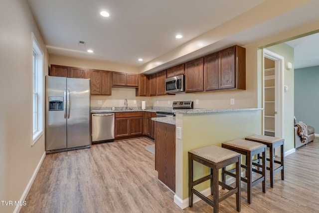 kitchen with kitchen peninsula, light hardwood / wood-style flooring, appliances with stainless steel finishes, a kitchen breakfast bar, and light stone counters