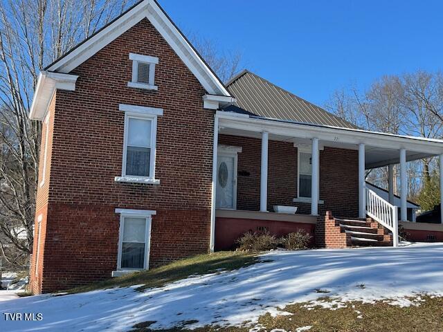 snow covered property with covered porch, brick siding, and metal roof