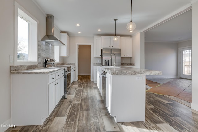 kitchen with an island with sink, white cabinets, hanging light fixtures, stainless steel appliances, and wall chimney range hood