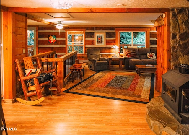 living room with a wood stove, wooden walls, beamed ceiling, ceiling fan, and light hardwood / wood-style floors