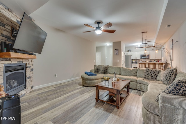 living room featuring baseboards, visible vents, a ceiling fan, a stone fireplace, and light wood-type flooring