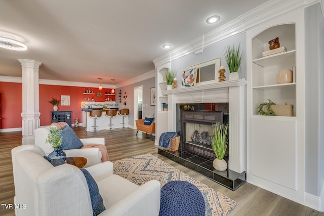 living room with crown molding, wood-type flooring, built in features, a tile fireplace, and decorative columns