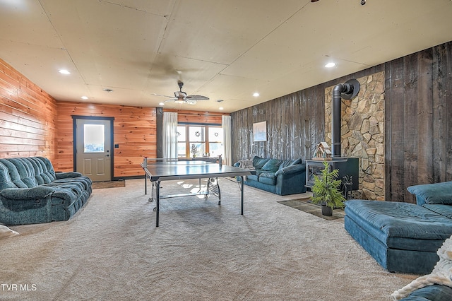 game room featuring ceiling fan, a wood stove, light carpet, and wood walls