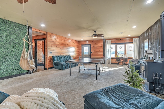 recreation room with wooden walls, carpet, ceiling fan, and a wood stove