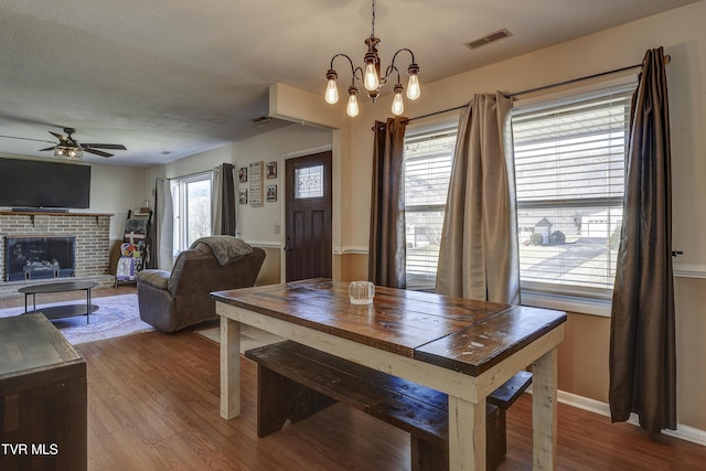 dining room featuring a brick fireplace, hardwood / wood-style flooring, ceiling fan with notable chandelier, and a textured ceiling