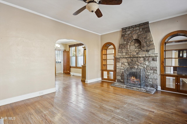 unfurnished living room featuring arched walkways, a stone fireplace, wood finished floors, baseboards, and ornamental molding