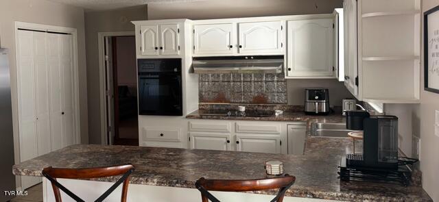 kitchen with a breakfast bar area, under cabinet range hood, white cabinets, dark stone counters, and black appliances