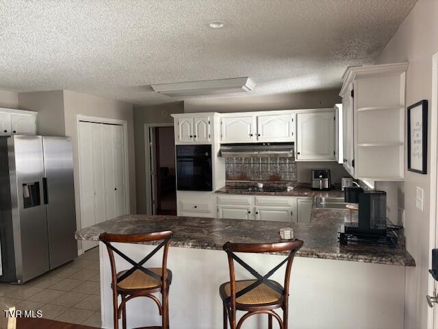 kitchen with light tile patterned floors, under cabinet range hood, a peninsula, white cabinets, and black appliances