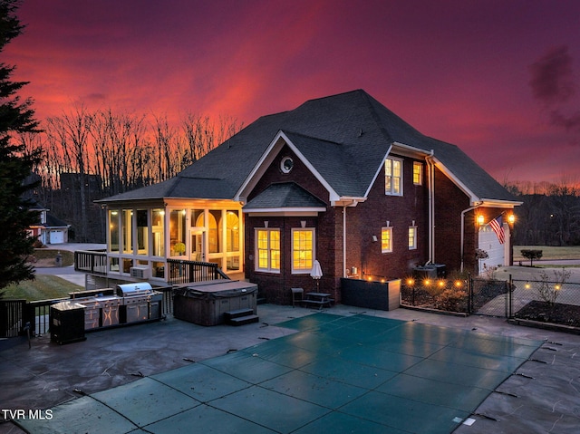 back of property at dusk with brick siding, a hot tub, a patio area, fence, and exterior kitchen