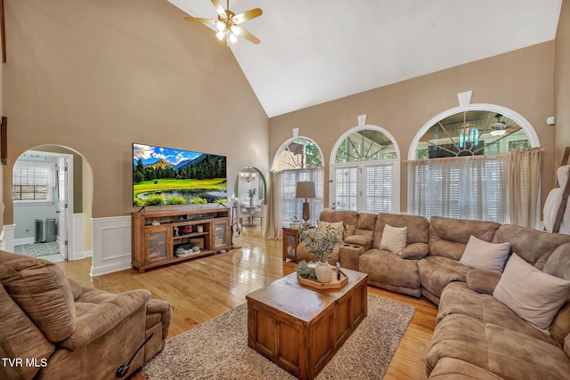 living area with arched walkways, a wainscoted wall, light wood-style floors, a ceiling fan, and high vaulted ceiling