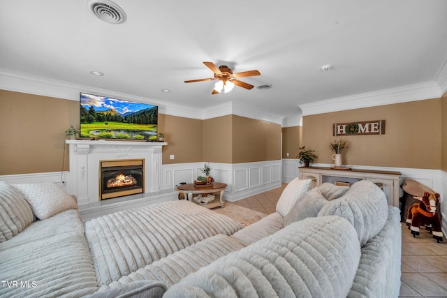 living room featuring light tile patterned floors, visible vents, wainscoting, a glass covered fireplace, and ornamental molding