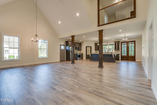 unfurnished living room with a chandelier, high vaulted ceiling, baseboards, light wood finished floors, and ornate columns