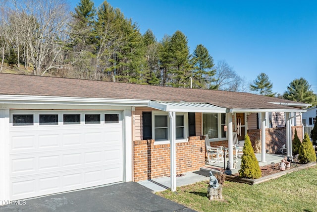 single story home with aphalt driveway, covered porch, a garage, a shingled roof, and brick siding