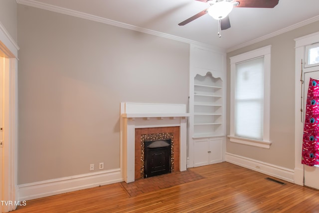 unfurnished living room featuring built in shelves, crown molding, a wood stove, light wood-type flooring, and ceiling fan