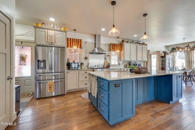 kitchen with stainless steel refrigerator with ice dispenser, wall chimney range hood, blue cabinets, black microwave, and hardwood / wood-style flooring