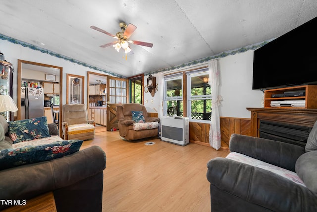 living room featuring vaulted ceiling, light wood-style floors, wainscoting, a ceiling fan, and a fireplace