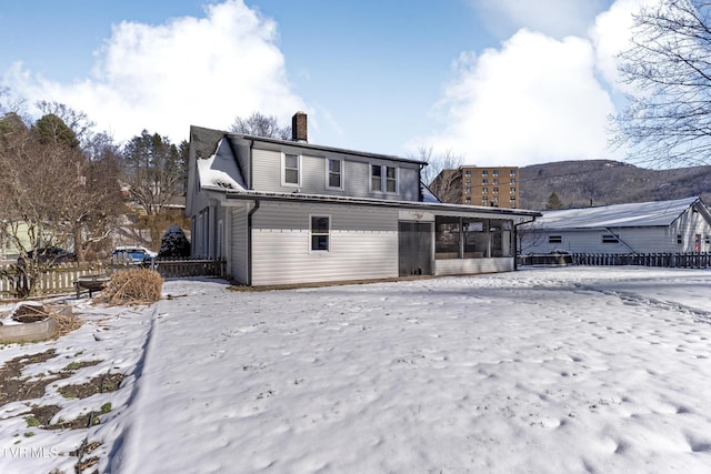 snow covered rear of property with a sunroom, a chimney, a mountain view, and fence