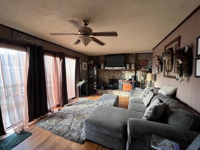 living area featuring a ceiling fan, a stone fireplace, a textured ceiling, and wood finished floors