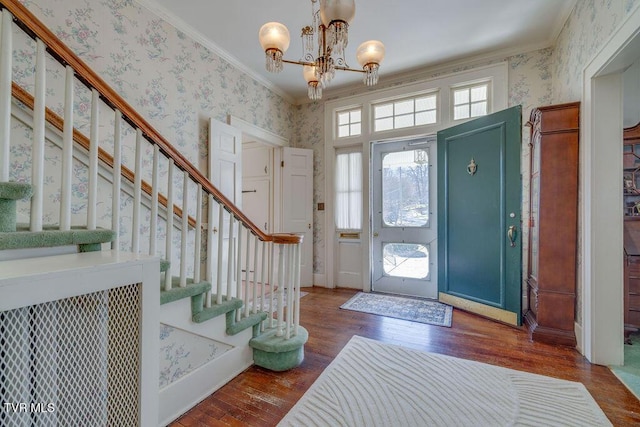 foyer entrance featuring dark wood-style flooring, stairway, an inviting chandelier, ornamental molding, and wallpapered walls