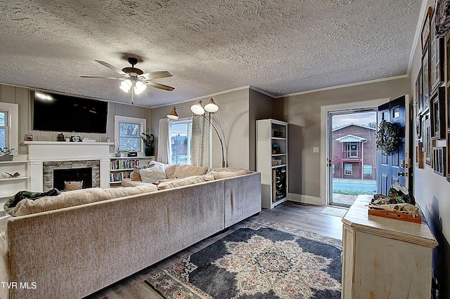 living room with dark wood-type flooring, ornamental molding, a ceiling fan, a stone fireplace, and a textured ceiling