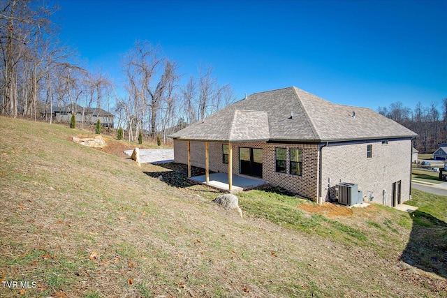 back of property with a patio, central AC, brick siding, a shingled roof, and a yard