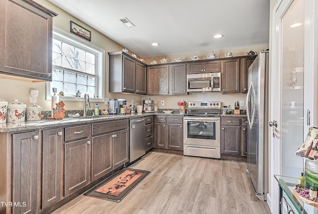 kitchen featuring stainless steel appliances, a sink, dark brown cabinets, light stone countertops, and light wood finished floors