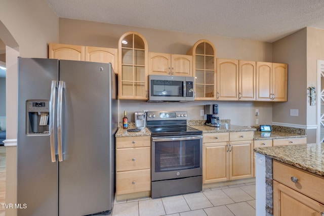 kitchen with light tile patterned floors, appliances with stainless steel finishes, light brown cabinets, and glass insert cabinets