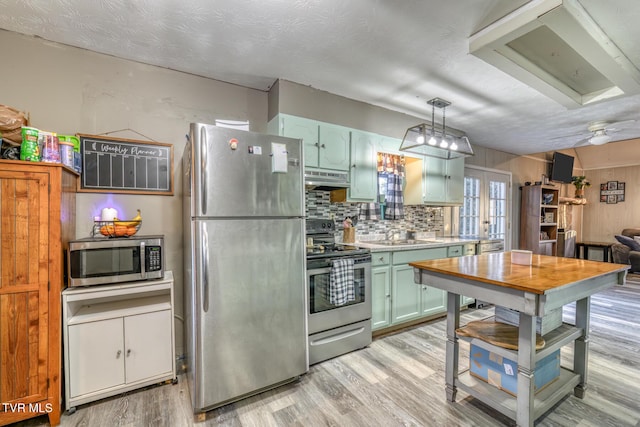 kitchen with light wood finished floors, appliances with stainless steel finishes, under cabinet range hood, green cabinets, and backsplash