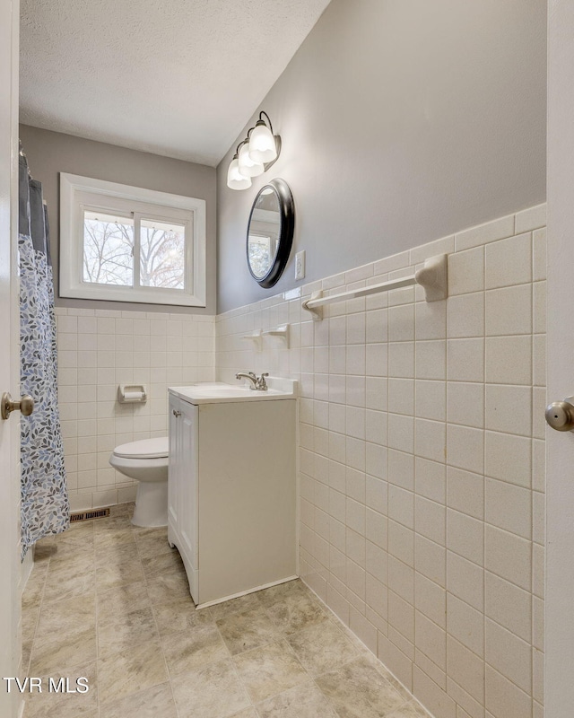 full bathroom featuring a textured ceiling, toilet, a wainscoted wall, vanity, and tile walls