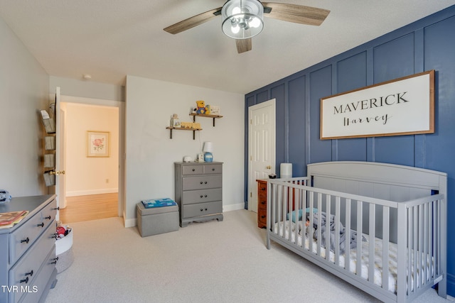 bedroom featuring baseboards, carpet flooring, a nursery area, a decorative wall, and a ceiling fan