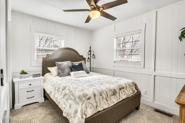 bedroom with a decorative wall, a ceiling fan, visible vents, and light colored carpet