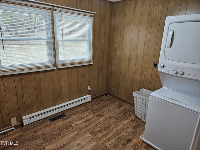 laundry room featuring stacked washer and dryer, laundry area, dark wood finished floors, wood walls, and a baseboard heating unit