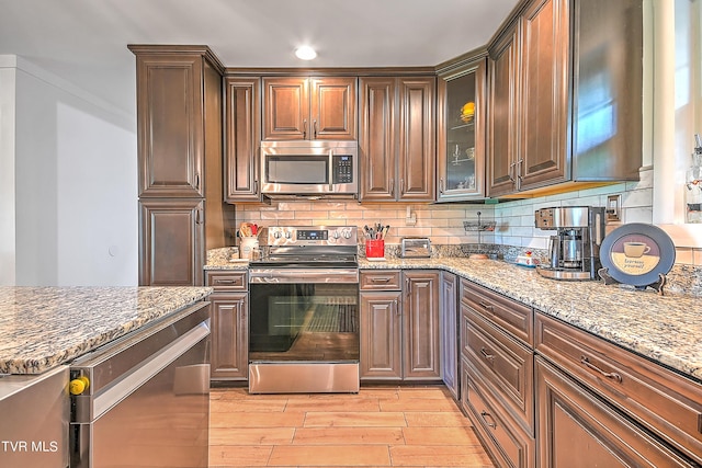 kitchen with light stone counters, light wood-style flooring, decorative backsplash, glass insert cabinets, and appliances with stainless steel finishes