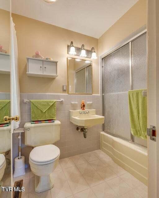 bathroom with a wainscoted wall, a sink, tile walls, toilet, and combined bath / shower with glass door