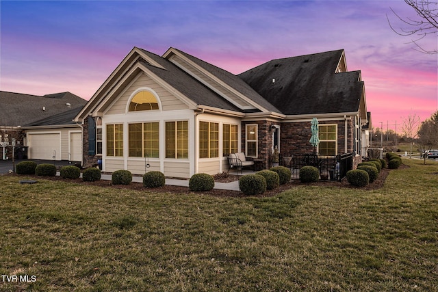 back of property at dusk with a garage, stone siding, a lawn, and a sunroom