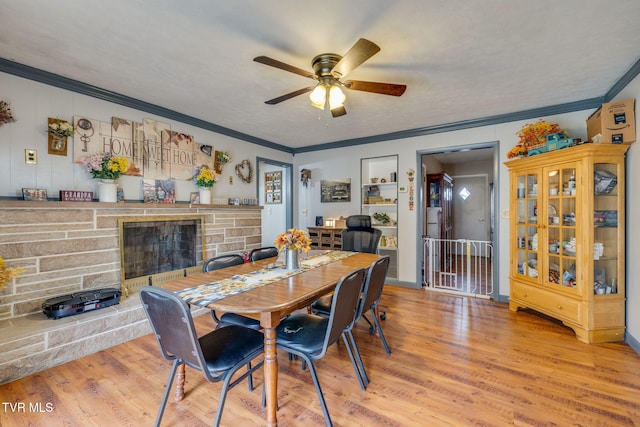 dining space featuring built in features, a ceiling fan, ornamental molding, a stone fireplace, and light wood-style floors