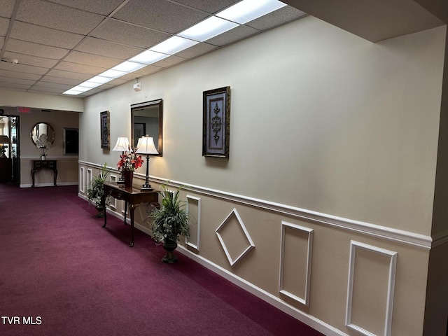 hall with a decorative wall, carpet flooring, a wainscoted wall, and a drop ceiling