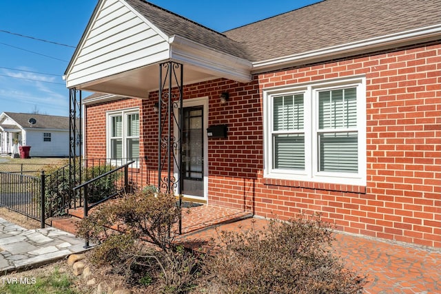 doorway to property featuring brick siding, roof with shingles, and fence