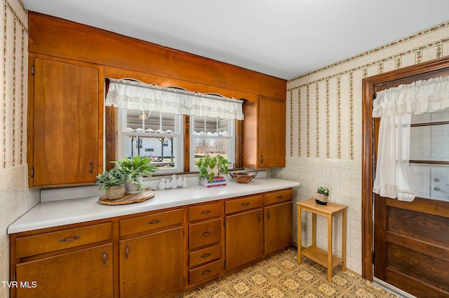 kitchen featuring brown cabinetry, wallpapered walls, tile walls, and light countertops