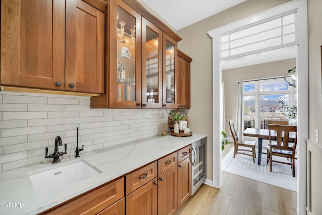 kitchen featuring light wood-type flooring, a sink, wine cooler, brown cabinetry, and light stone countertops