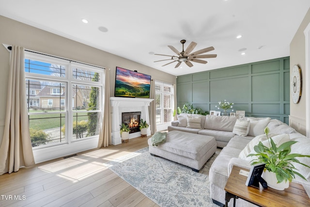 living area with visible vents, a ceiling fan, a glass covered fireplace, a decorative wall, and light wood finished floors