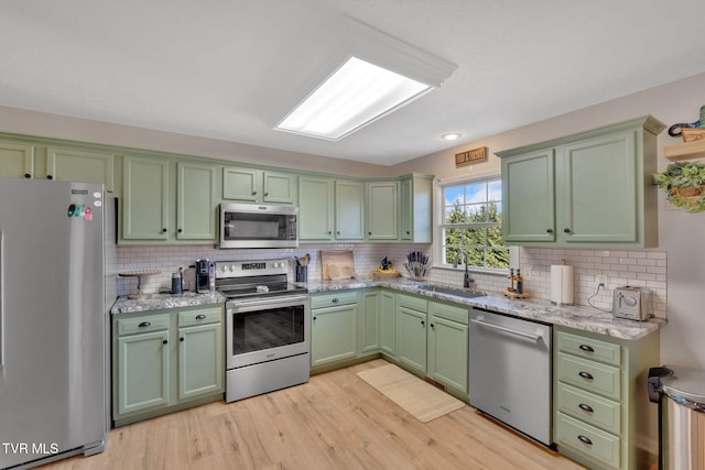 kitchen with a sink, backsplash, stainless steel appliances, light wood finished floors, and green cabinetry