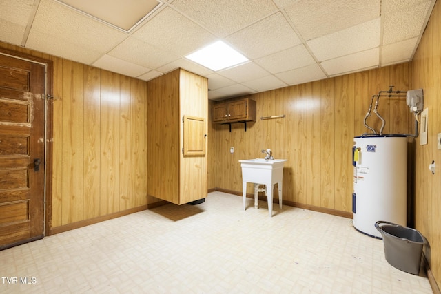 basement with light floors, a sink, a paneled ceiling, wood walls, and electric water heater